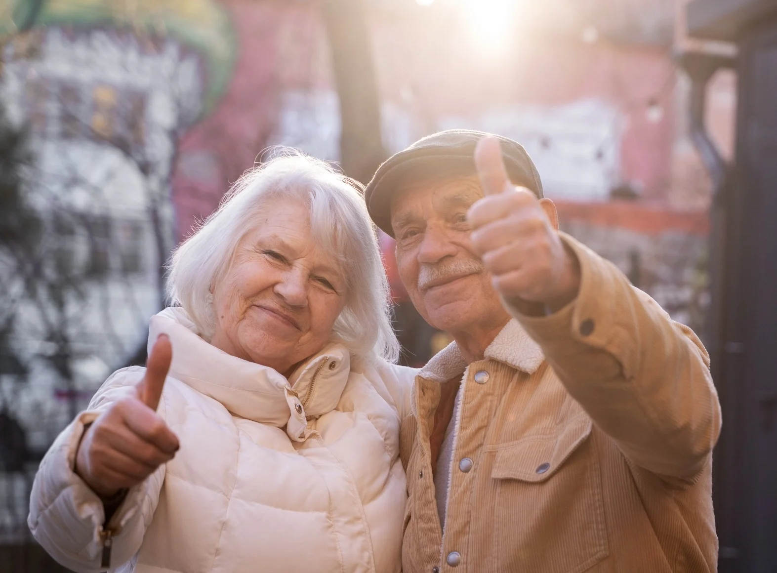 Coppia di anziani sorridenti che mostrano il pollice in su all’aperto in un contesto urbano con luce naturale e atmosfera positiva